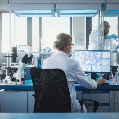 Medical Laboratory with Team of Scientists Working. Microbiologist is Using Personal Computer, Screen Shows High-Tech Concept for DNA research. High-Tech Biotechnology Research. Back View Shot